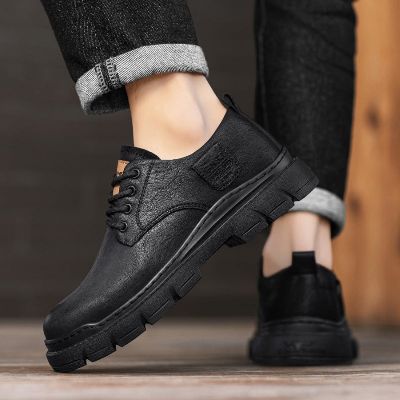 Black leather shoes worn with rolled-up jeans on a wooden floor.