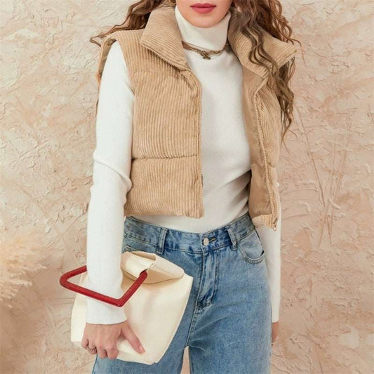 Person wearing a beige corduroy vest over a white turtleneck and blue jeans, holding a beige handbag with a red handle against a beige background.