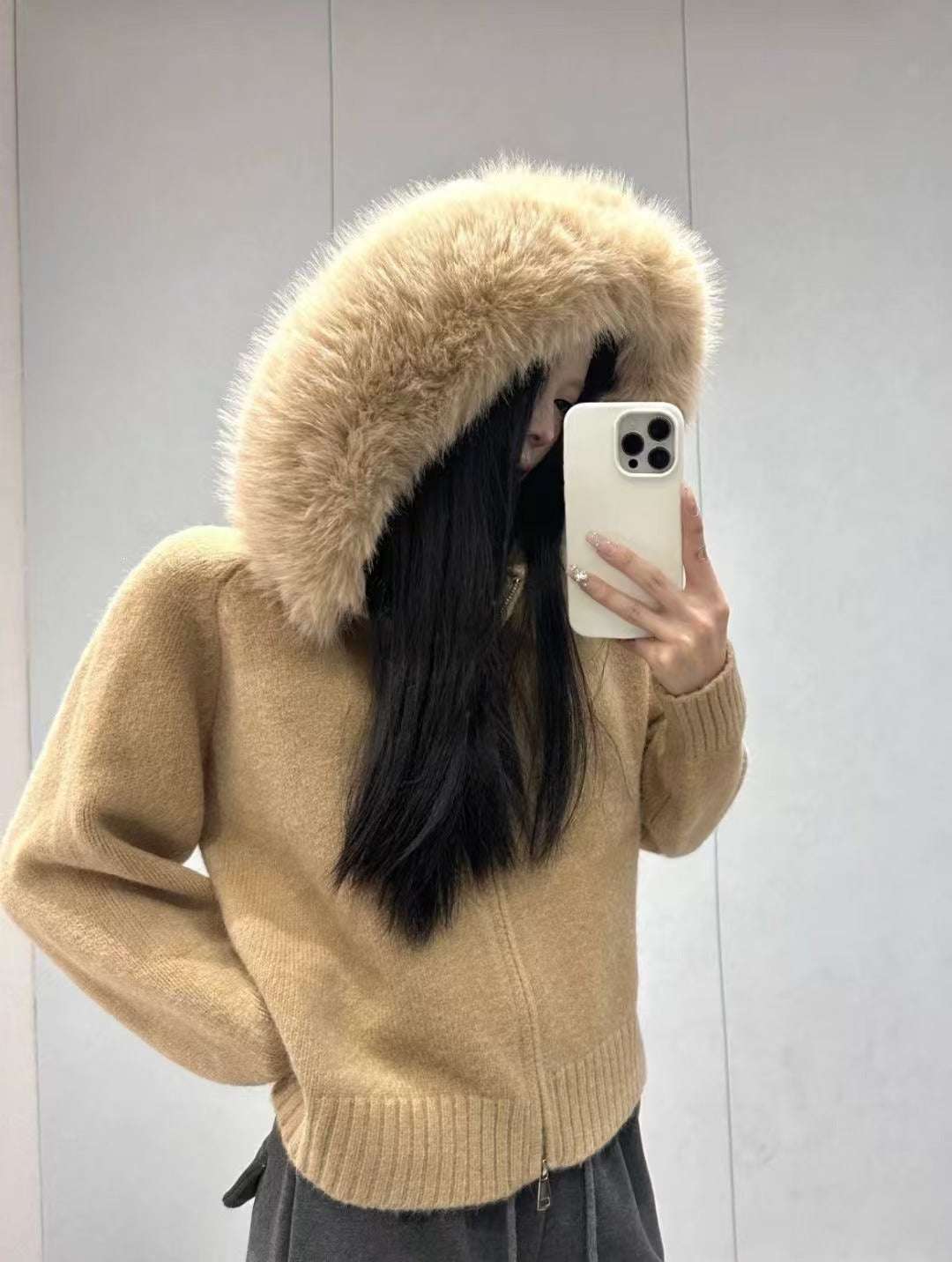 Person wearing a beige sweater with a fur-lined hood, taking a mirror selfie.