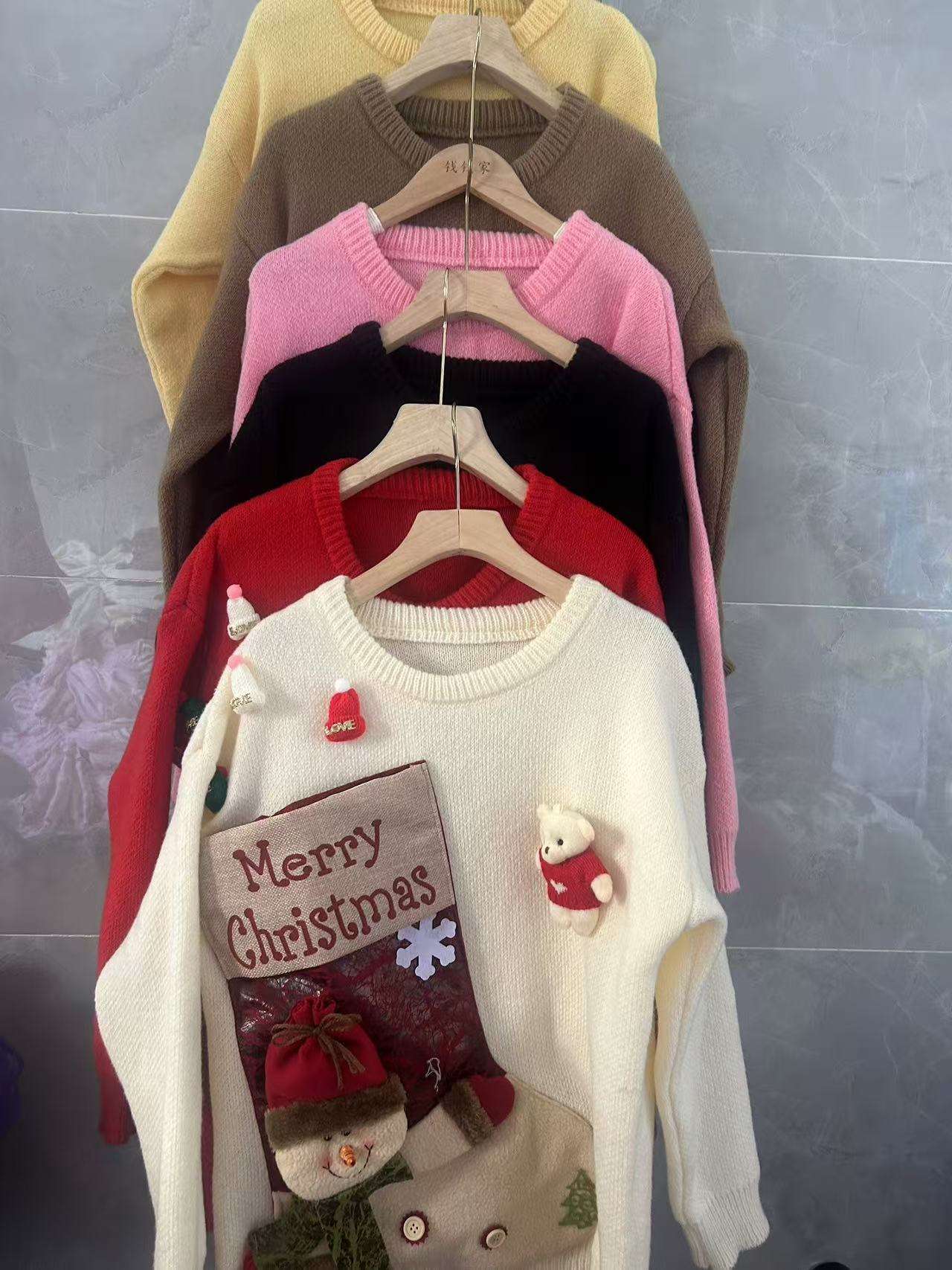 Stack of children's sweaters with 'Merry Christmas' design on a hanger against a gray wall.