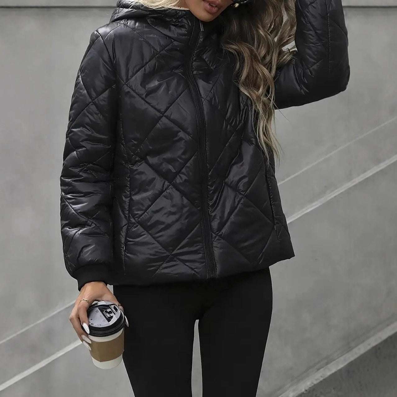 Person wearing a black quilted jacket holding a coffee cup against a gray background