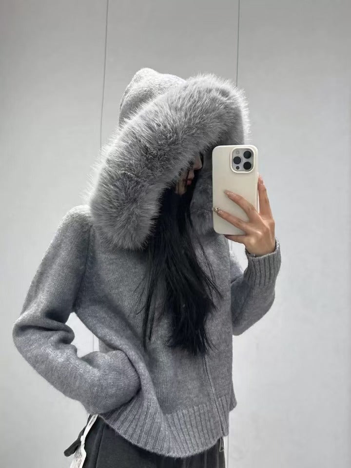 Person wearing a gray sweater with a large fur hood, holding a phone.