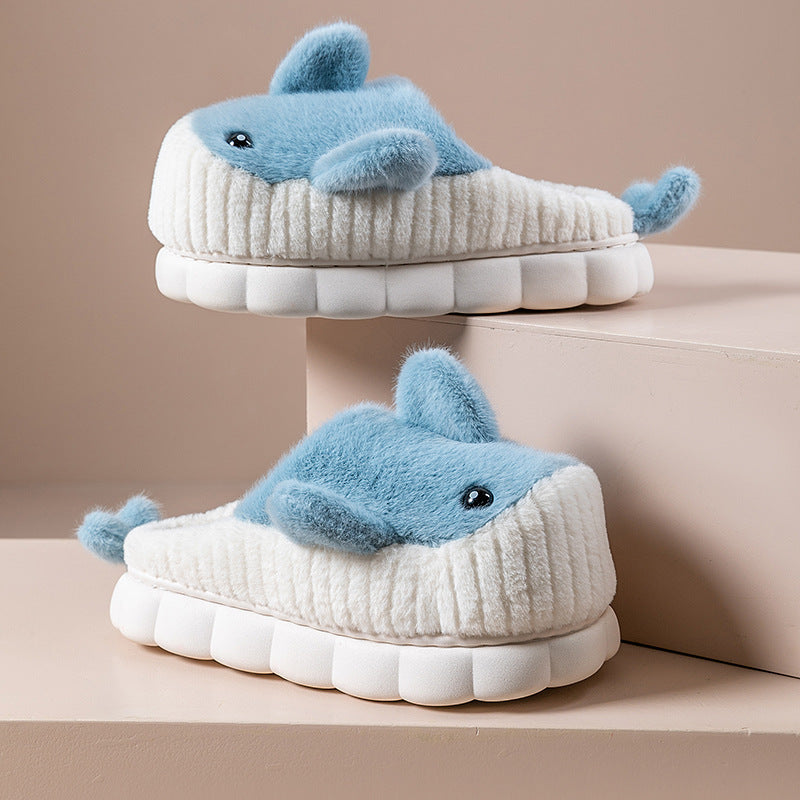 A pair of women's home slippers designed to resemble a cute shark, with a thick white sole and blue plush upper, set against a beige background.