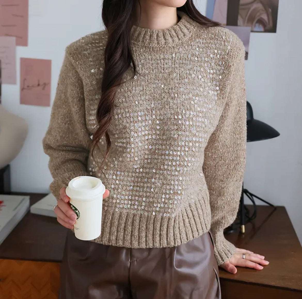 Person wearing a sparkly beige sweater holding a white cup indoors.