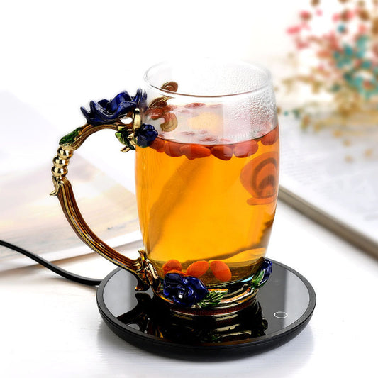 Glass mug with floral handle on a black heating pad, filled with a hot beverage.