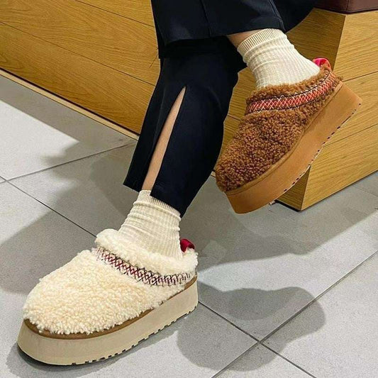 A pair of fluffy closed-toe slippers with thick soles worn by a person sitting, predominantly in beige and brown colors.