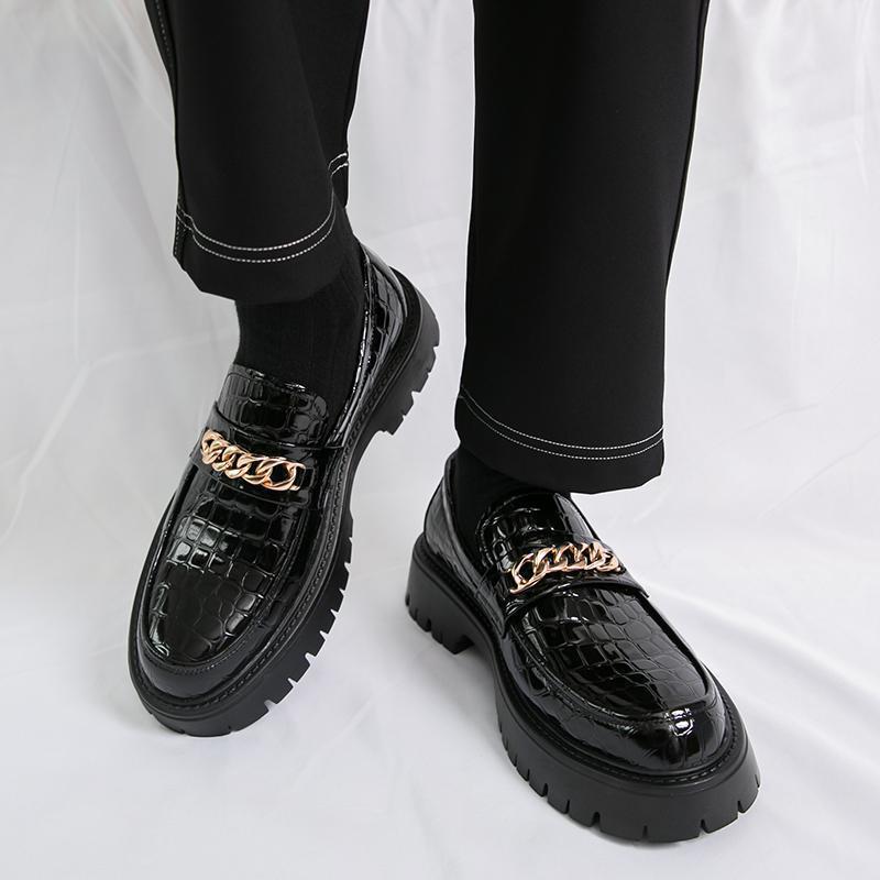 Black crocodile-patterned shoes with gold chain details worn by a person on a white background