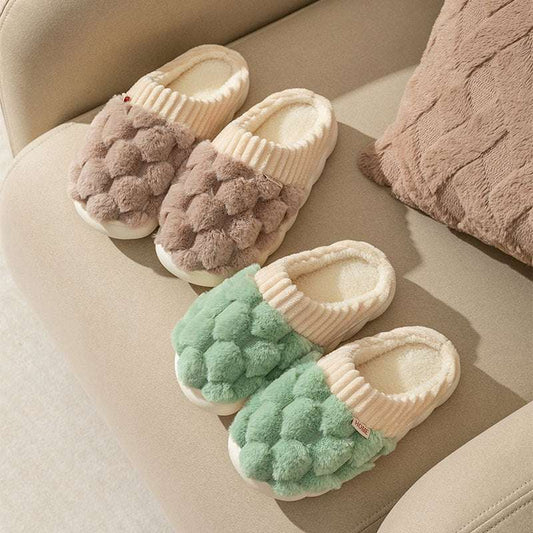 A pair of brown and a pair of green plush slippers with non-slip sole, placed on a sofa.