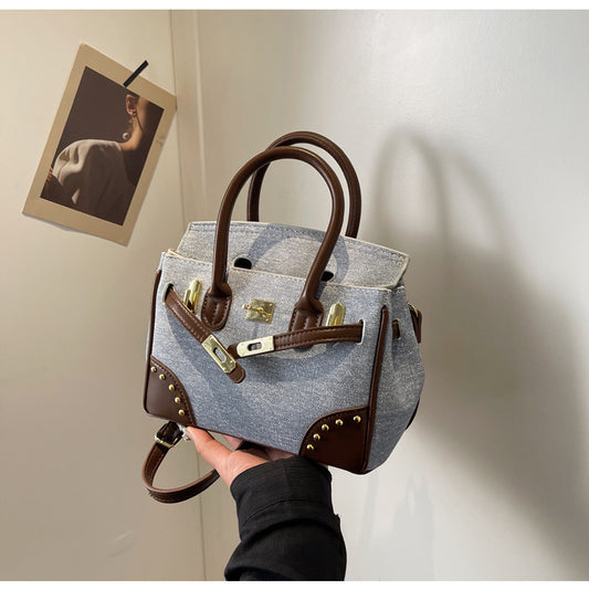 A hand holding a portable commuter bag with a blue denim-like pattern and brown leather straps and details.