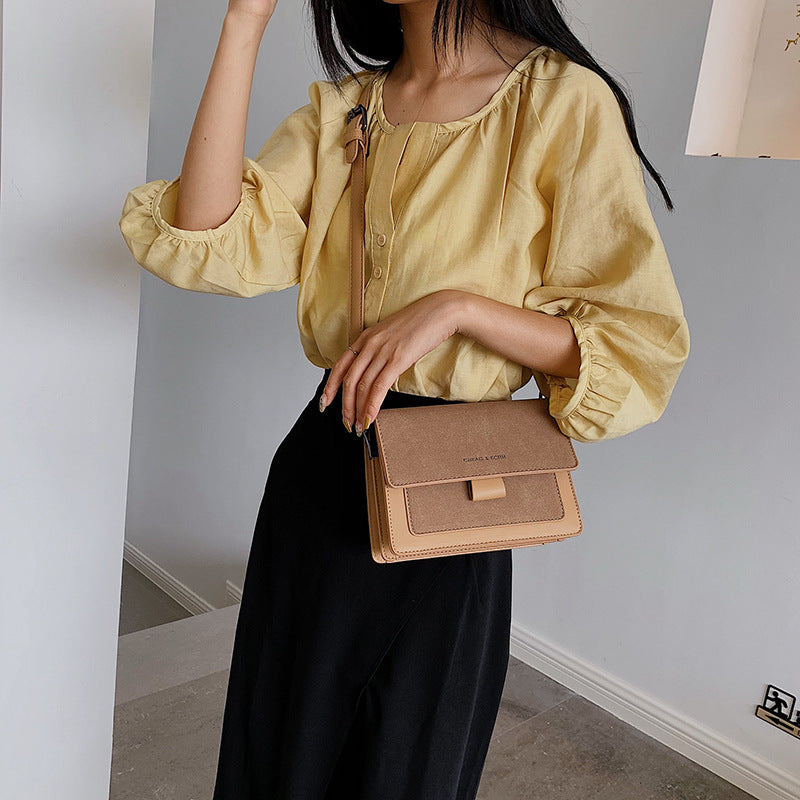 A person wearing a yellow top and black skirt holding a small square-shaped beige bag with a contrast color design and a matte finish.