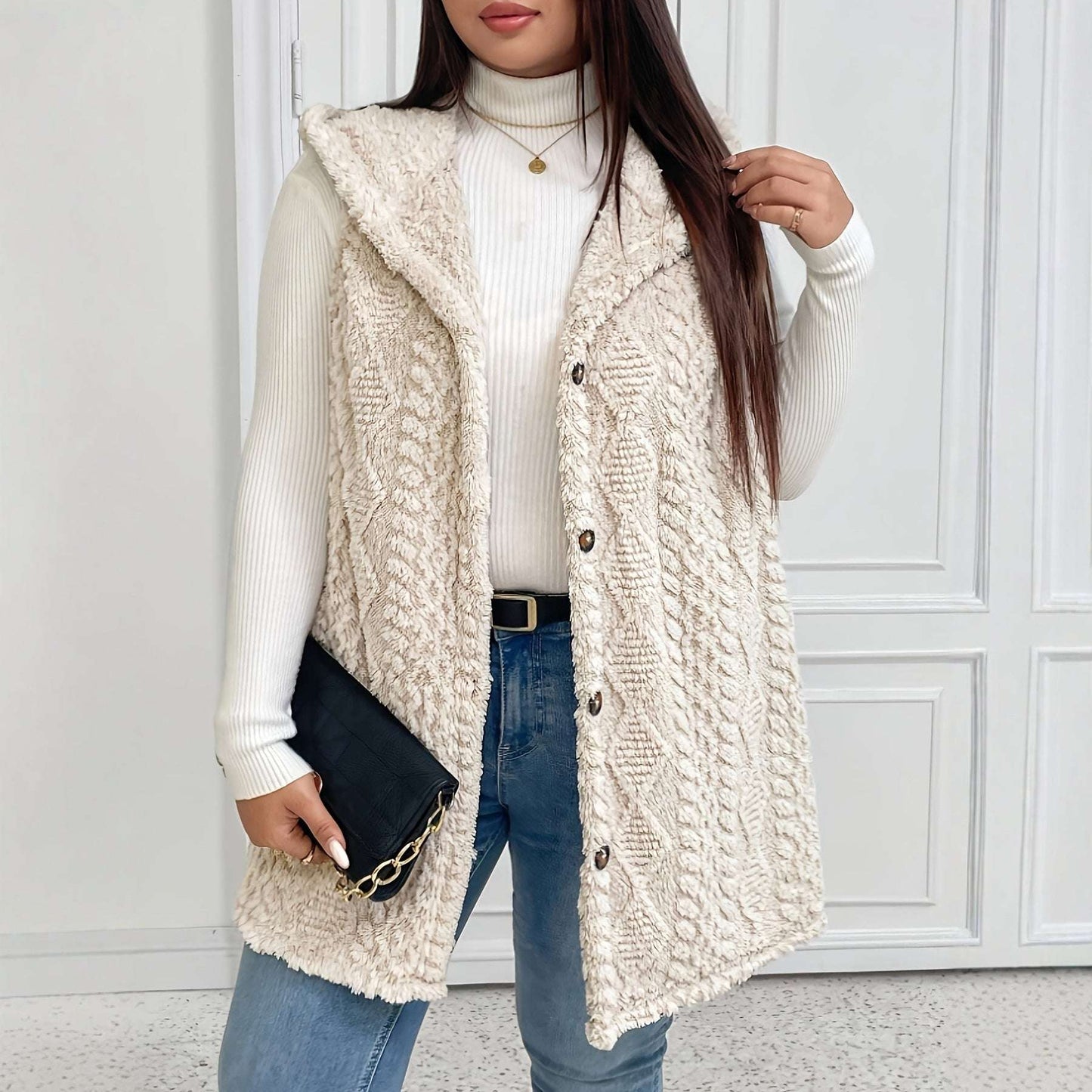 Woman wearing a cream knitted vest over a white turtleneck and blue jeans, holding a black clutch.