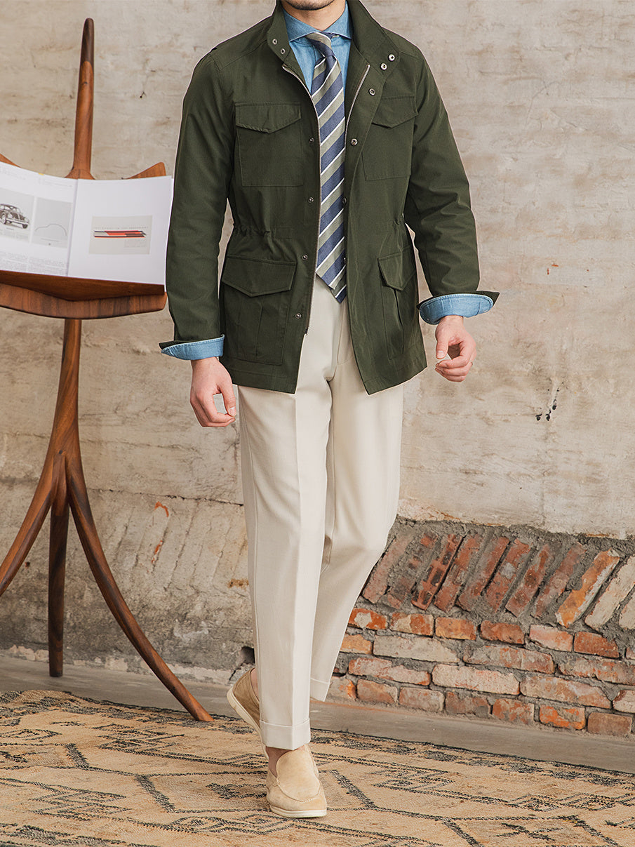Man wearing a green jacket, striped tie, and beige pants standing in front of a brick wall.