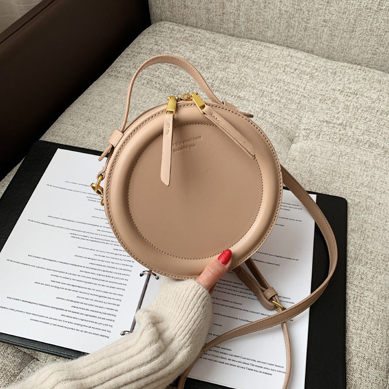 Light tan round zip-top crossbody bag with gold-tone hardware and adjustable shoulder strap