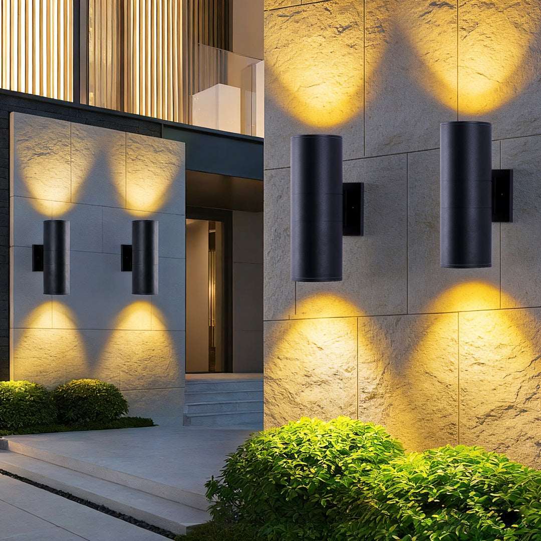 Matte black cylinder outdoor wall lights on stone wall with warm up-lighting and plants