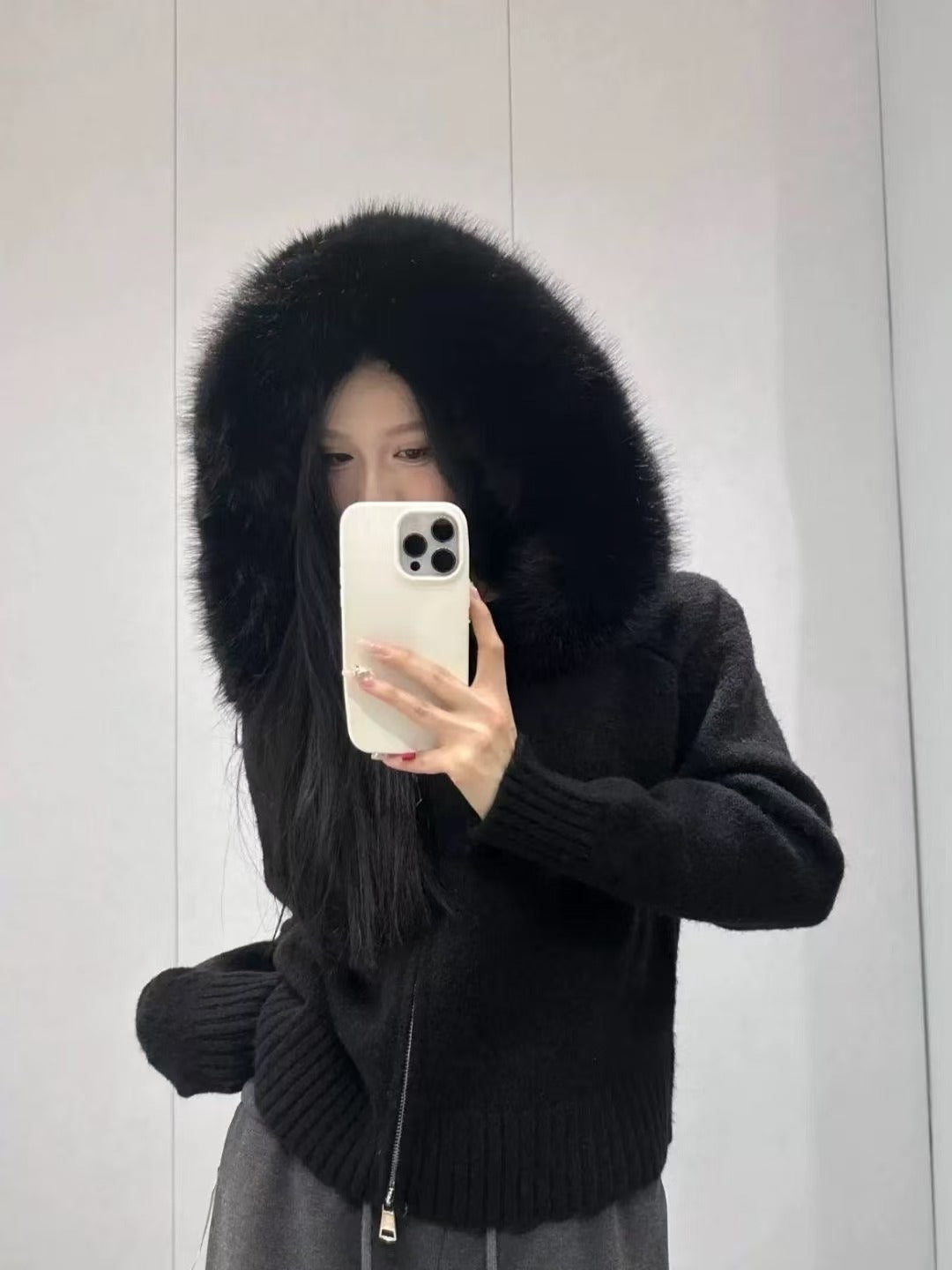 Person wearing a black coat with a fur-lined hood, holding a phone against a plain background.