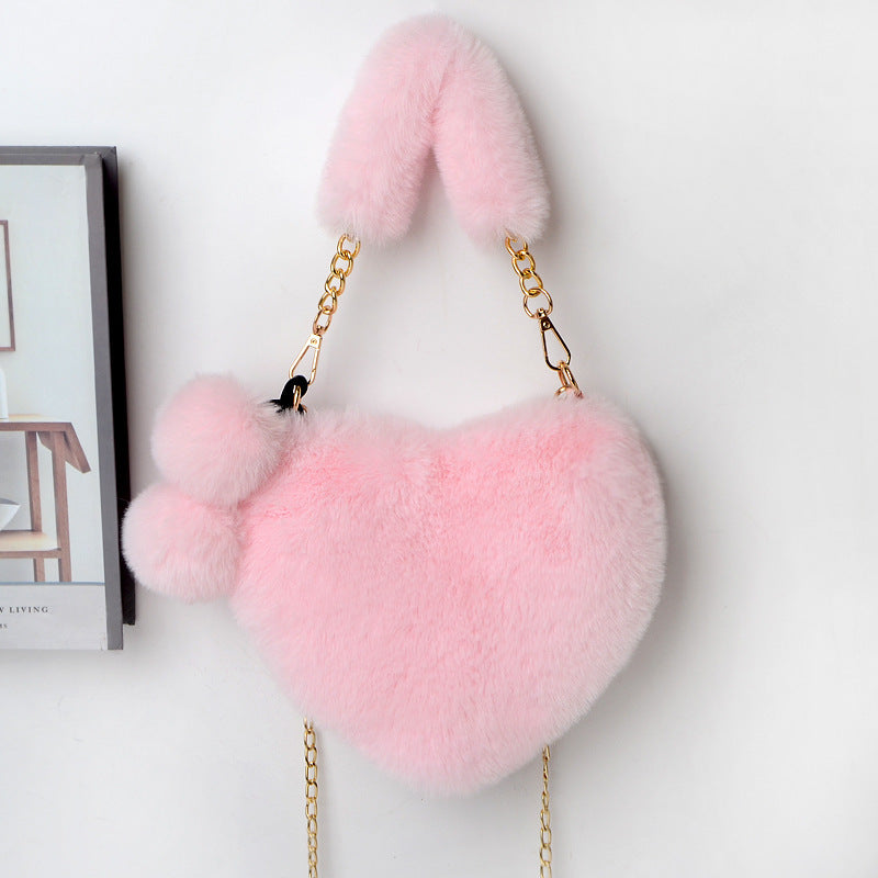Light pink plush heart shoulder bag with gold chain and fuzzy pom-pom charm.