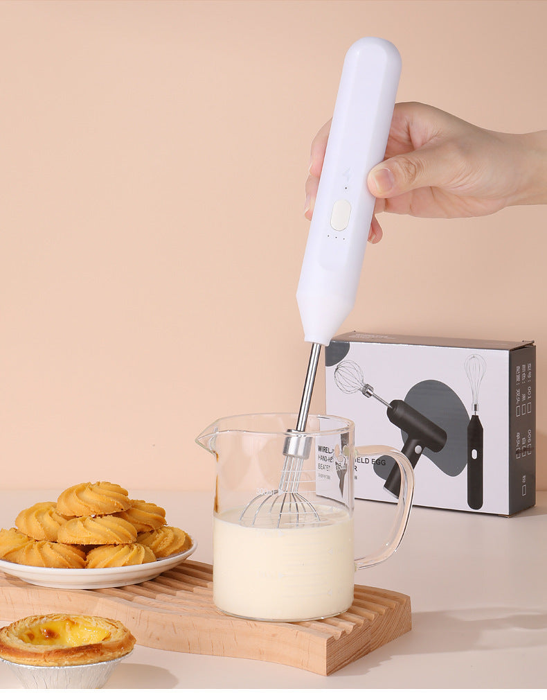 Hand holding a white whisk over a glass pitcher with milk, with cookies and a box in the background.