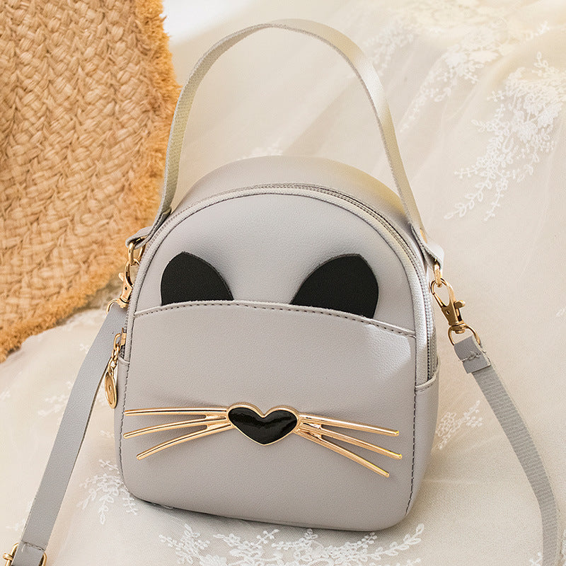 Light grey mini cat-face backpack with black ears, gold heart whisker bar, and strap