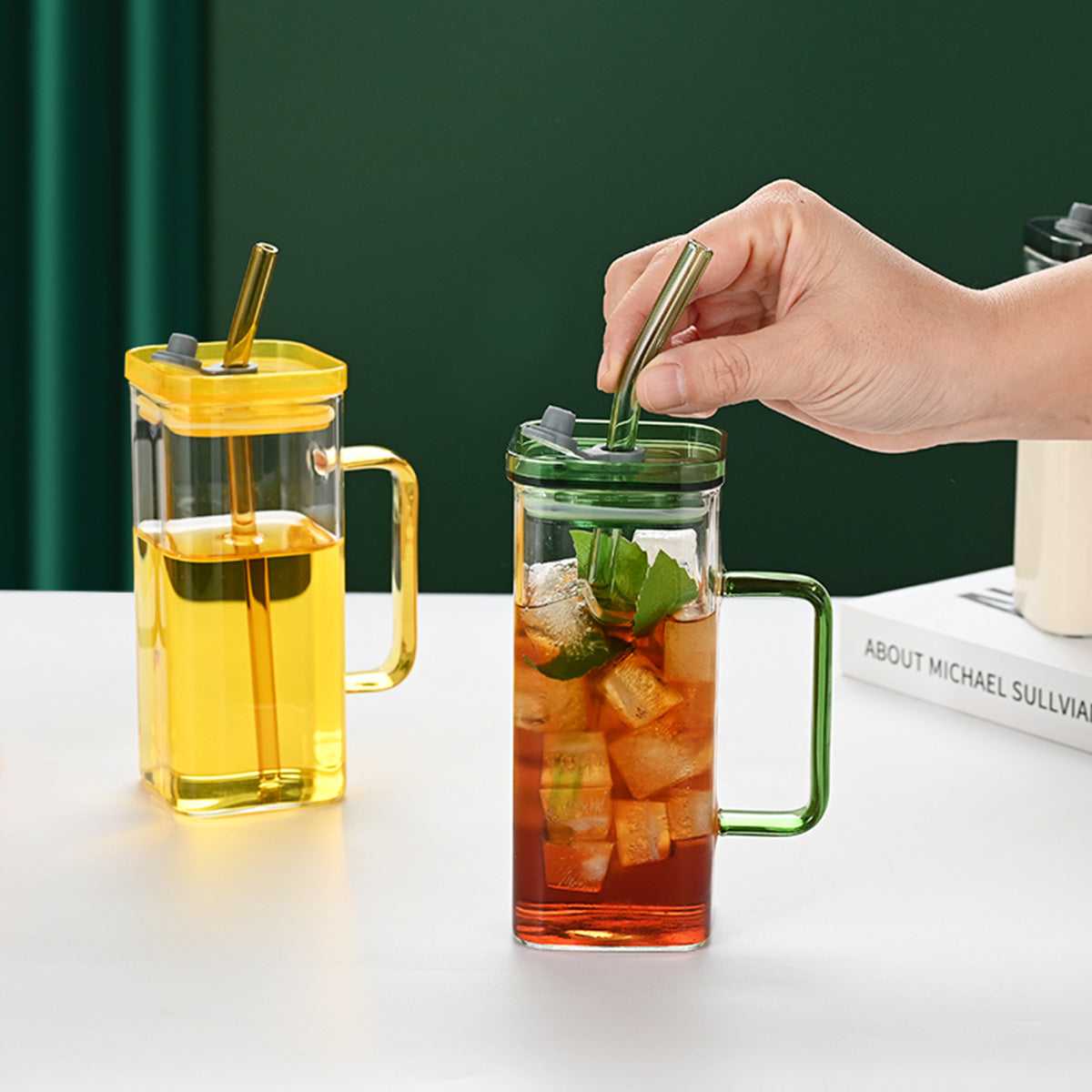 Clear square glass cup with green lid and handle, with another yellow-lidded cup nearby