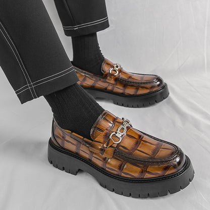 Brown crocodile-patterned loafers with black soles worn by a person on a light gray background