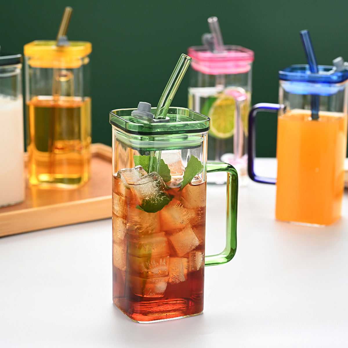 Square glass cup with green lid and straw, plus orange and blue cups