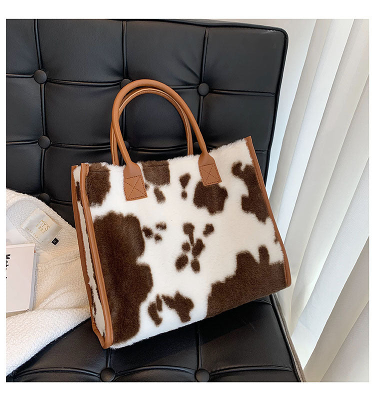 Cow-print plush tote bag with brown faux-leather handles and square front tabs