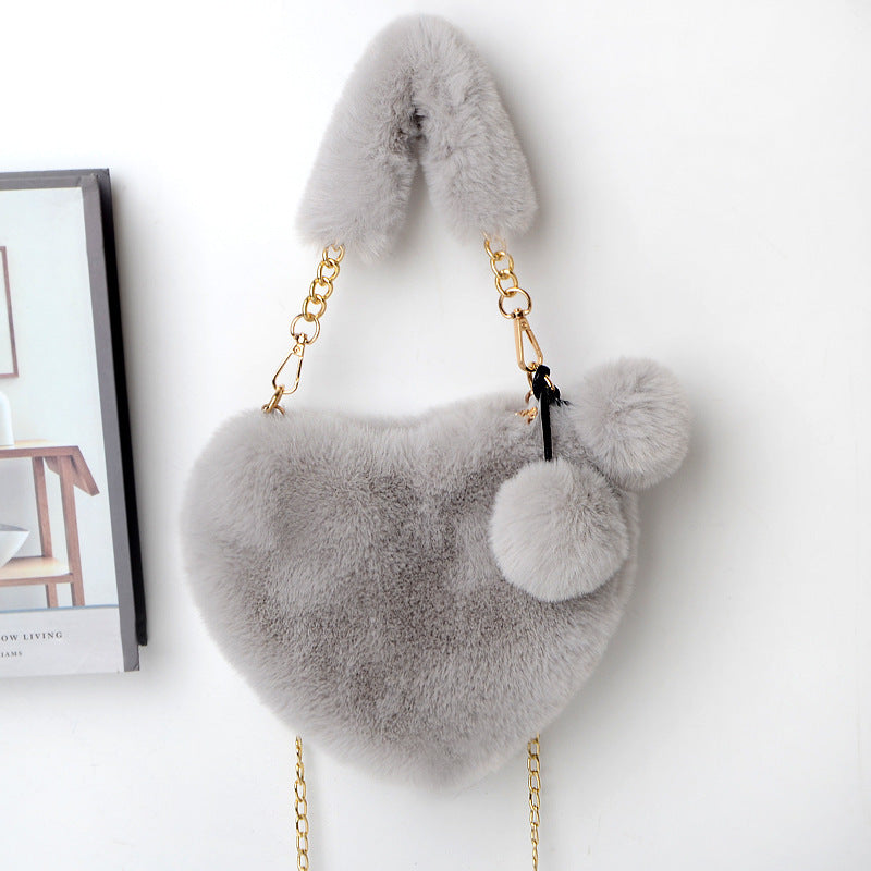 Light gray plush heart shoulder bag with gold chain and side pom-pom details.