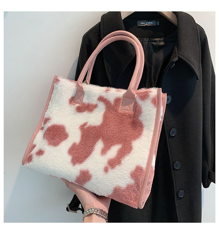 Cow-print plush tote bag with pink-brown tones, pink handles, and square logo tabs