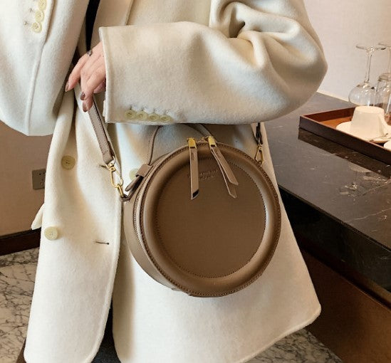 Taupe round zip-top crossbody bag with gold-tone hardware and adjustable shoulder strap