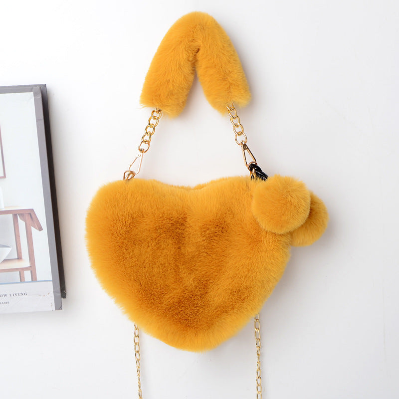 Mustard yellow plush heart shoulder bag with gold chain and fuzzy pom-pom charm.