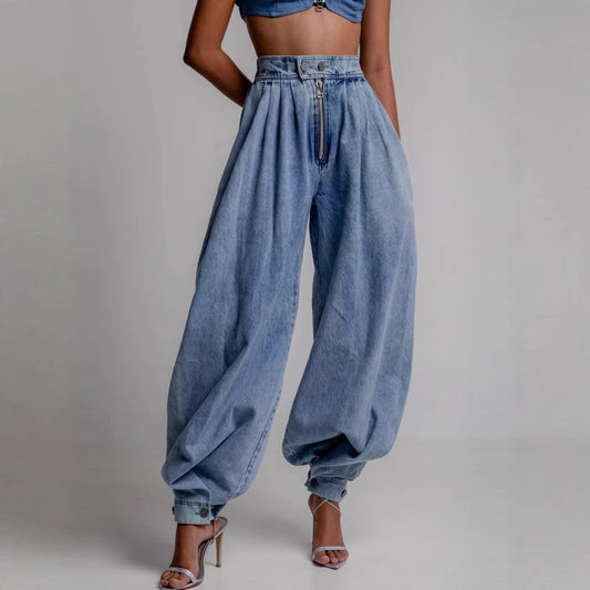 A woman modeling loose-fitting harem-style denim jeans with a high-waisted cut, visible zipper fly, and a tapered ankle finish.
