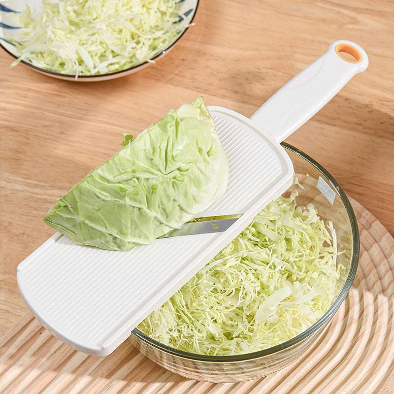 Cabbage being shredded with a manual slicer onto a glass bowl on a wooden surface.