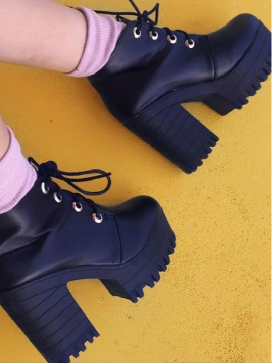 A pair of black high-heel chunky heel ankle boots with lace-up closure on a yellow background.