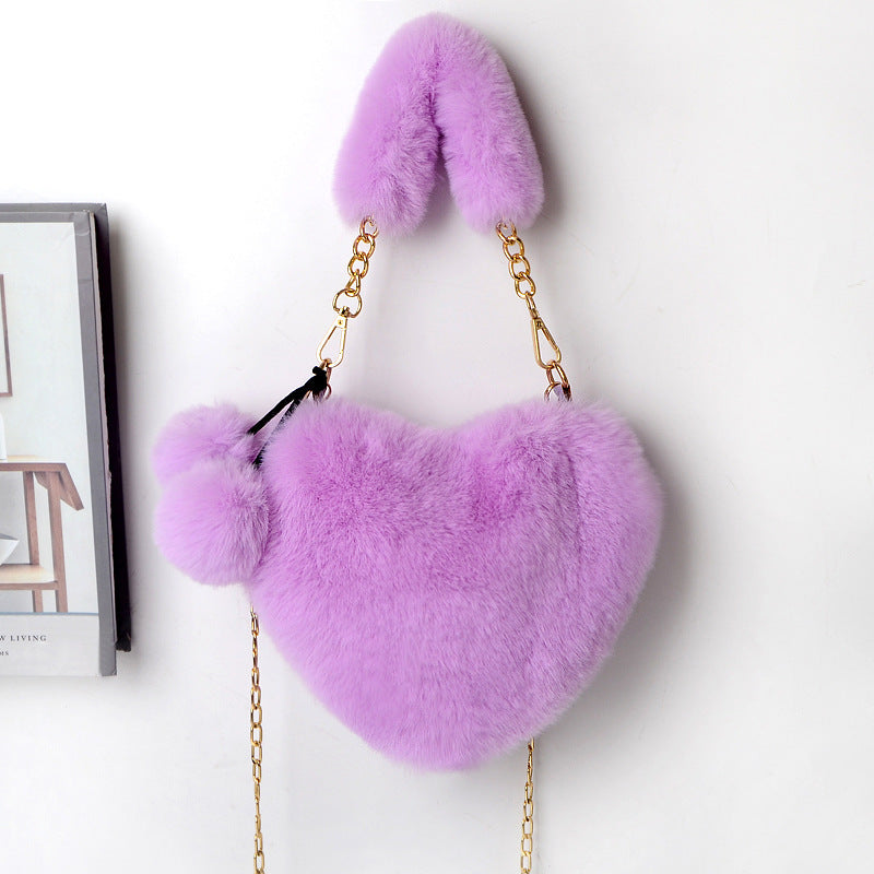 Heart-shaped plush handbag with pom-pom accents and gold chain strap, lavender purple