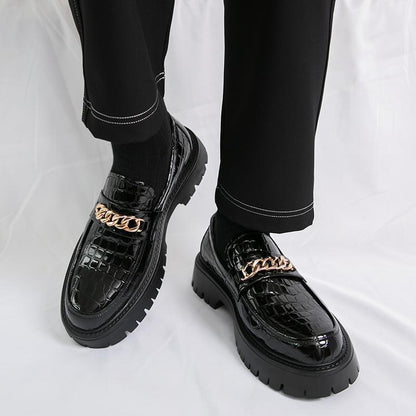 Black crocodile-patterned shoes with gold chain details worn by a person on a white background