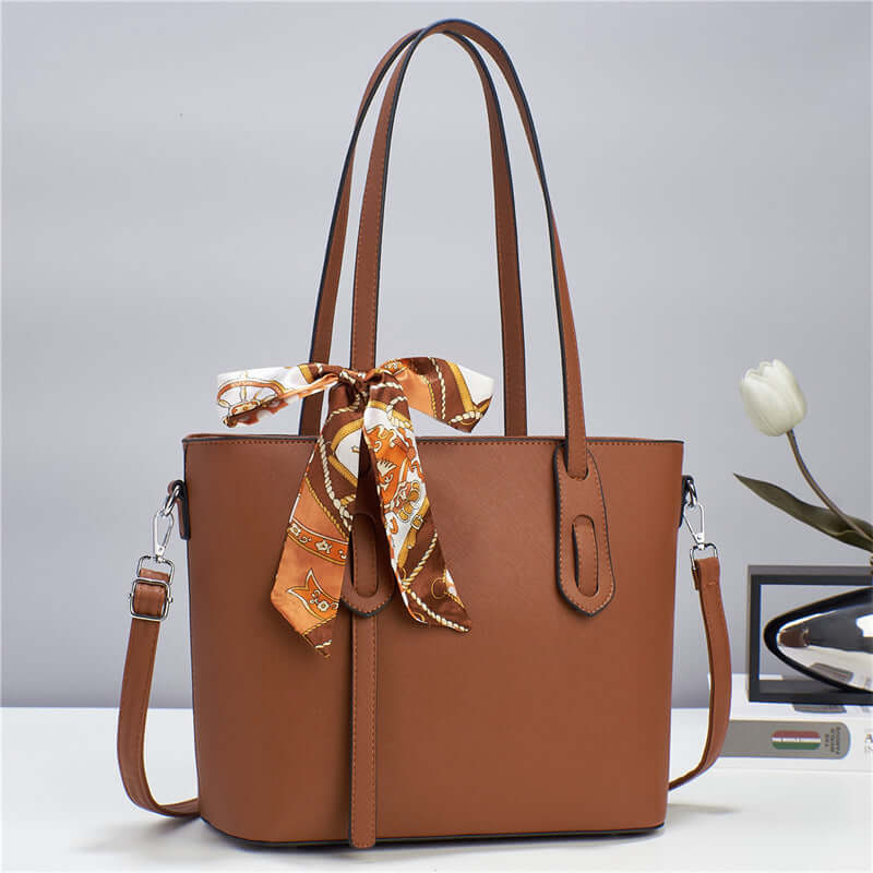 Brown tote handbag with top handles, front zipper pocket, and tied patterned scarf at top.