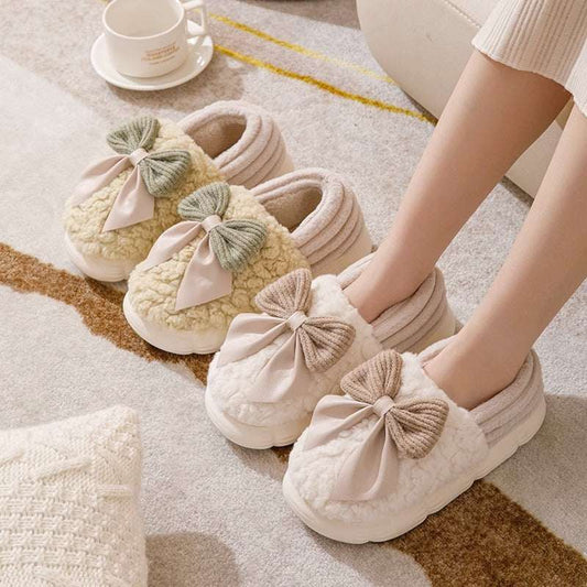 A pair of beige fluffy slippers with a big bow-knot on the top, displayed in a home setting with a cup of coffee and a knit sweater in the background.