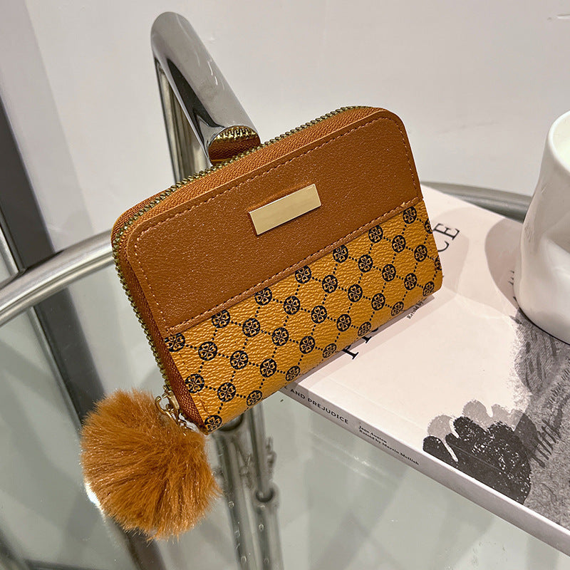 Brown patterned short wallet with tan top flap, gold clasp, and pom-pom