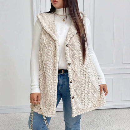 Person wearing a beige knitted vest over a white sweater and blue jeans against a white wall.