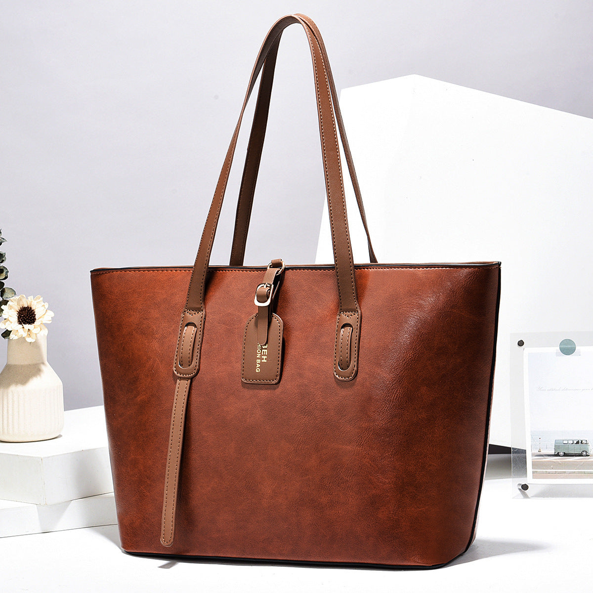 Brown leather tote handbag with two long handles and front vertical strap accents.