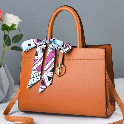 Large orange crossbody tote with brown handles, colorful ribbon scarf, and metal clasp charm