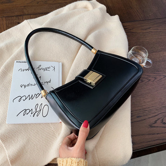 A woman sitting with crossed legs, wearing a beige sweater, black pants, and black shoes, carrying a black shoulder bag with a gold clasp.