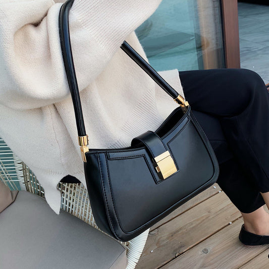 A woman sitting with crossed legs, wearing a beige sweater, black pants, and black shoes, carrying a black shoulder bag with a gold clasp.