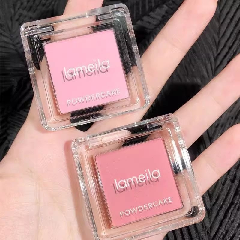 Two pink powder cakes with 'lameila' branding held in a hand against a black textured background.