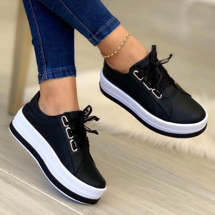 Black sneakers with white soles worn by a person on a wooden floor.