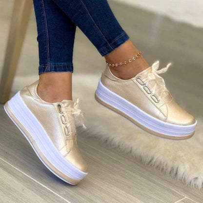 Beige sneakers with white soles worn with blue jeans on a light wooden floor.
